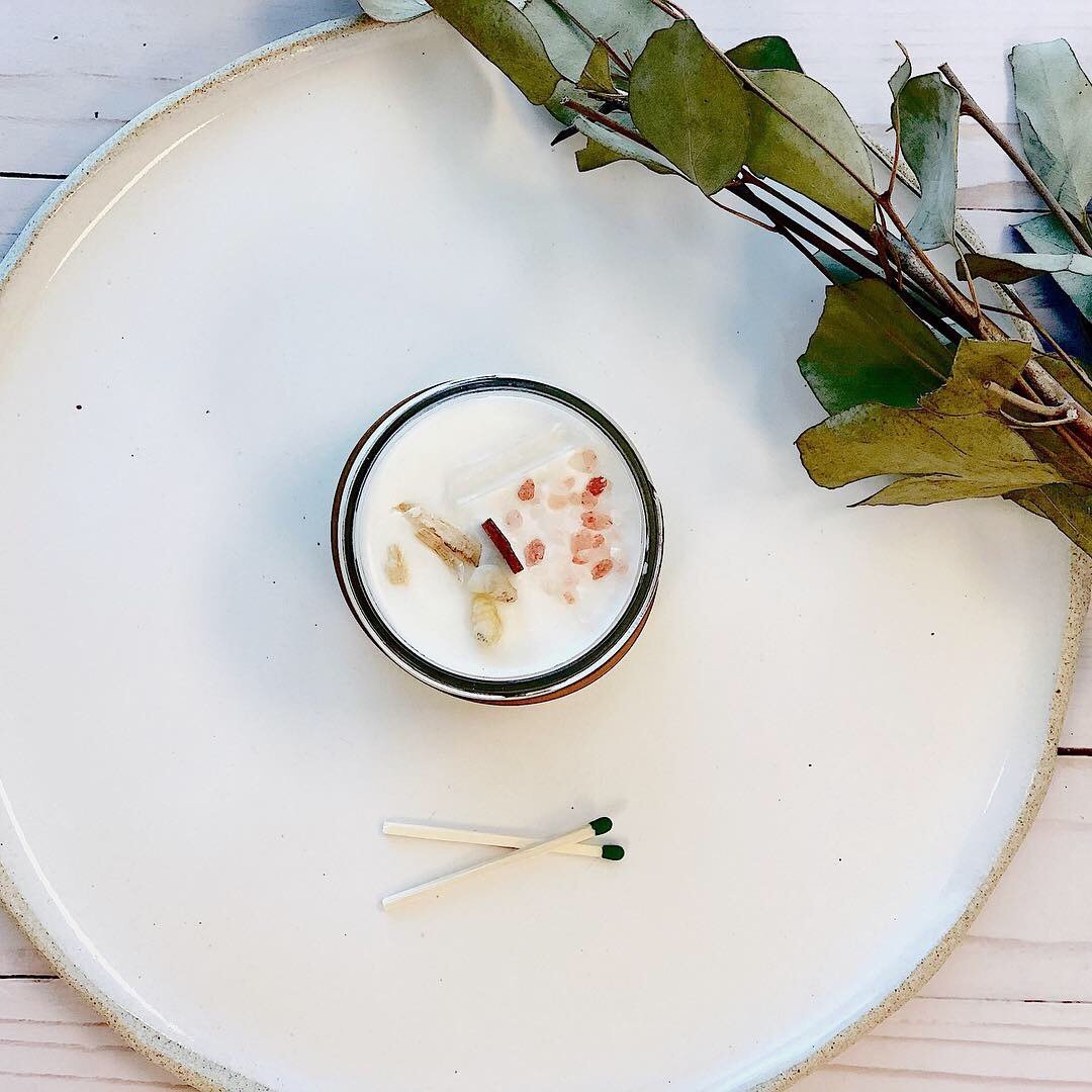Space clearing intention candle with herbs and crystals on a white plate beside matchsticks.