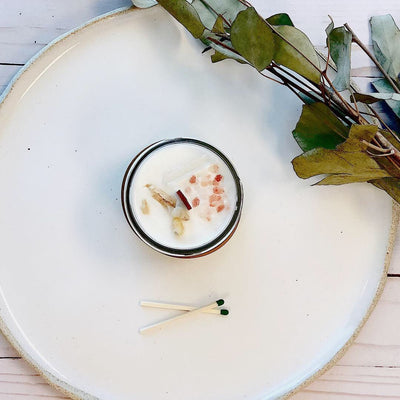 Space clearing intention candle with herbs and crystals on a white plate beside matchsticks.